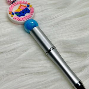 May include: A silver pen with a blue ballpoint tip and a decorative top. The top features a white and pink circular charm with the words "Emotional Dumpster Fire" and a cartoon dumpster on fire.