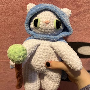 Puede incluir: Un gato de peluche tejido a ganchillo en color blanco, con una capa azul con capucha y un pequeño bastón con una punta esférica verde. El gato tiene grandes ojos negros con iris verde claro y una pequeña nariz rosa, hecho de hilo suave.