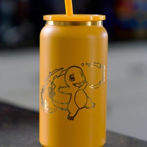 Puede incluir: Vaso de color amarillo mostaza con tapa y pajita a juego. El vaso presenta un gráfico de color dorado de un personaje de dibujos animados. El diseño está grabado en la superficie del vaso. El vaso es cilíndrico y parece ser de metal.