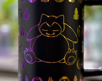 May include: Black stainless steel travel mug with a wraparound design featuring a gold-colored Snorlax character and other colorful graphics. The rim of the mug has a rainbow gradient. A black handle is attached to the side.