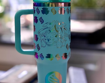 May include: A light blue insulated tumbler with a teal handle and lid. The tumbler features a holographic design of a cartoon character and decorative accents. The bottom of the tumbler has a holographic emblem. The tumbler is on a black stand.