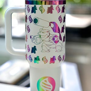 May include: White travel mug with a handle, featuring a colorful Blastoise design. The mug has a white lid and handle, with a rainbow-colored band at the top and a circular logo at the bottom. The Blastoise graphic is outlined in rainbow colors.
