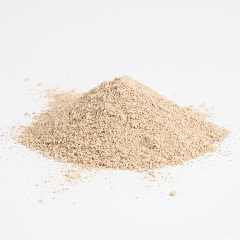 May include: A pile of light brown powder sits against a white background. The powder has a fine, granular texture, with small particles visible throughout. The shape is roughly conical, with a slightly uneven surface. The color is a muted beige.