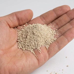May include: A close-up shot of a hand holding a pile of light brown, granular powder. The powder appears to be finely ground and has a slightly textured appearance. The background is plain white.