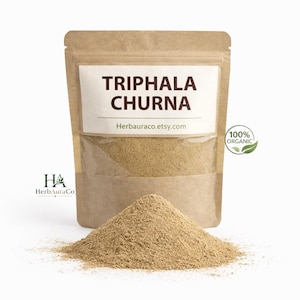 May include: A brown kraft paper pouch containing Triphala Churna powder, with a white label reading Triphala Churna and Herbauraco.etsy.com. A small circular 100% Organic logo with a green leaf is visible, and a pile of the light brown powder sits in front.