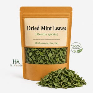May include: A brown stand-up pouch containing dried green mint leaves, labeled Dried Mint Leaves [Mentha spicata] with the URL Herbauraco.etsy.com. A circular 100% Organic seal is visible, and a pile of loose dried mint leaves sits in front.
