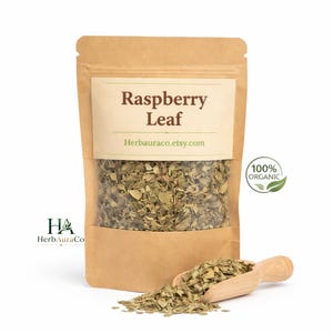 May include: A brown paper bag with a clear window filled with dried raspberry leaves. A wooden scoop holds a pile of the leaves, with more scattered in front. The label reads "Raspberry Leaf" and "Herbauraco.etsy.com". A green leaf graphic indicates "100% Organic".