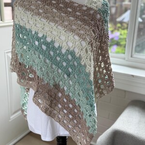 May include: A crochet poncho featuring a diagonal striped pattern in shades of tan, sage green, and cream. The garment is constructed with an open, airy stitch design that creates a repeating square grid texture throughout the entire piece.