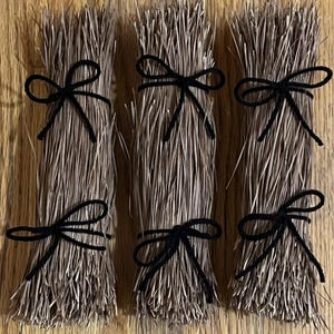 Peut inclure: Trois bottes d'aiguilles de pin séchées brun clair, chacune fixée par deux nœuds en cordelette noire attachés en haut et en bas. Les bottes sont disposées verticalement, mettant en valeur la texture fine et effilée du matériau naturel.