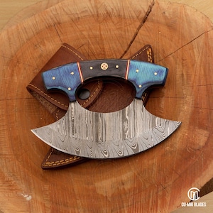 May include: A Damascus steel karambit knife with a blue and black handle, resting on a wooden surface. The handle has gold accents and is paired with a brown leather sheath. The blade has a curved, crescent shape.
