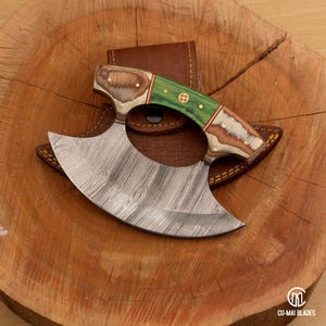 Op de afbeelding: Een Damascus stalen hakmes met een gebogen lemmet en een groen- en houtkleurig handvat. Het mes ligt op een houten oppervlak met een bruine leren schede. De handgreep heeft een decoratief messing accent.