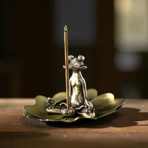 Puede incluir: Un porta incienso de rana de bronce con una base de hoja de loto. La rana está en posición sentada, sosteniendo un palito de incienso marrón. La hoja es de color verde apagado.