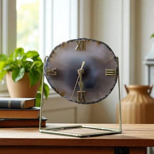 May include: A decorative table clock with a round face made from a polished, grey and white stone. The clock features gold Roman numerals and hands, set on a gold metal stand. A unique home decor item.