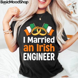 May include: Black t-shirt with the text "I Married an Irish ENGINEER" in white and orange. The design includes two gold wedding rings and Irish flags. The shirt is a casual wear item.