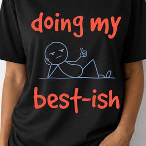 Pode incluir: Camiseta preta com a frase "doing my best-ish" em letras vermelhas. Apresenta também uma figura de palito azul com o polegar para cima. É uma peça de roupa casual.