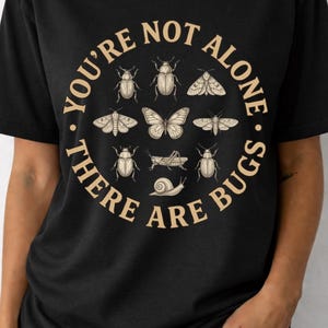 May include: Black t-shirt with a beige circular design featuring insect illustrations. The text "YOU'RE NOT ALONE. THERE ARE BUGS" surrounds the insects. The shirt is made of a soft material and is suitable for casual wear.