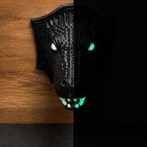 Könnte beinhalten: Eine schwarze Wandplakette in Form eines Drachenkopfes mit einer texturierten, schuppenartigen Oberfläche. Der Drache hat leuchtend hellgrüne Augen und Zähne, die sich vor dem dunklen, kontrastreichen Hintergrund deutlich abheben.