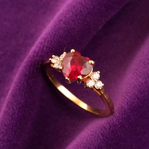 May include: A gold ring with a heart-shaped, red gemstone as the centerpiece. The ring is adorned with smaller, clear gemstones on either side of the heart. The ring is set against a purple velvet background.