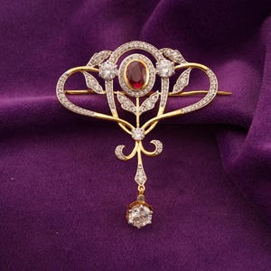 Puede incluir: Broche dorado con un rubí ovalado en el centro, rodeado de piedras preciosas claras y delicados diseños de hojas. Un único diamante redondo cuelga debajo. El broche se encuentra sobre un fondo púrpura.