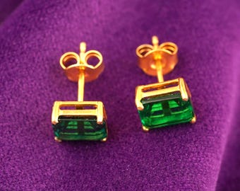 Pode incluir: Um par de brincos de botão dourados com pedras preciosas retangulares verde esmeralda. Os brincos são cravejados em uma moldura dourada e possuem fecho de pressão. São exibidos em uma superfície roxa.