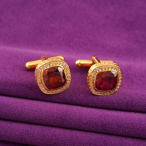 May include: A pair of gold cufflinks featuring square, deep red gemstones. Each cufflink is framed by a halo of small, clear crystals. The cufflinks are displayed on a purple fabric background.