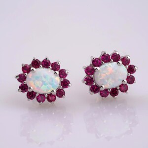 May include: A pair of silver-toned stud earrings. Each earring features an oval, iridescent opal center stone surrounded by a halo of small, round, deep red gemstones. The earrings are set against a white background.