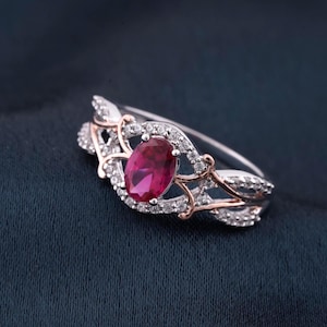 May include: A silver ring with a large oval ruby center stone, surrounded by small diamonds. The ring has a decorative design with rose gold accents, set against a dark blue background.