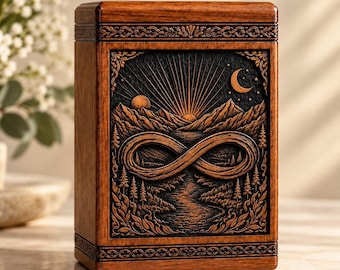 May include: A rectangular wooden cremation urn featuring a dark, recessed central panel with a carved relief of a mountain landscape, a river, a crescent moon, a sun, and an infinity symbol. The top and bottom edges have decorative borders.