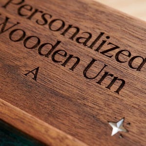 May include: Close-up of a wooden urn with the words "Personalized Wooden Urn" engraved on the lid. A star-shaped cutout is visible on the right side. The urn is made of dark brown wood.