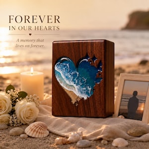 May include: A wooden urn with a heart-shaped cutout revealing a beach scene with blue and white waves. The text "FOREVER IN OUR HEARTS" and "A memory that lives on forever" are displayed. A lit candle, flowers, and a framed photo are also present.