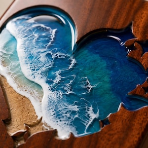 May include: Wooden box with a heart-shaped cutout filled with a resin ocean scene. The ocean design features shades of blue and white, with a sandy beach effect. The box is made of dark wood.
