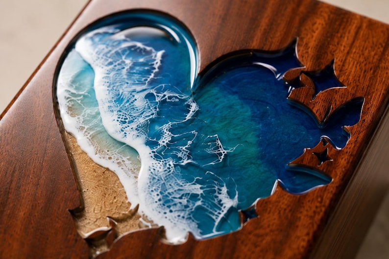 May include: Wooden box with a heart-shaped cutout filled with a resin ocean scene. The ocean design features shades of blue and white, with a sandy beach effect. The box is made of dark wood.