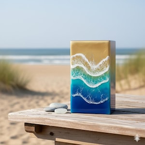 Puede incluir: Una pieza de arte rectangular con una escena de playa. La parte superior es dorada, pasando a olas blancas y espumosas sobre agua turquesa y azul oscuro. Dos pequeñas piedras grises y un trozo de vidrio marino se encuentran sobre una superficie de madera.