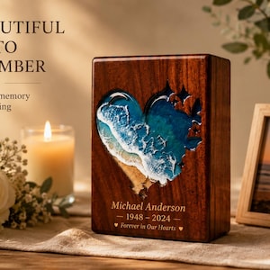 May include: A wooden memorial urn with a heart-shaped ocean scene carved into the front. The urn has the name "Michael Anderson" and dates "1948-2024" engraved on it. A framed photo and a lit candle are also in the image.