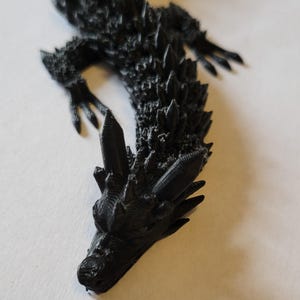 May include: A black, 3D-printed dragon figurine with a textured surface and spiky details. The dragon is lying down on a white surface, with its head and claws visible. The figurine is approximately 12 inches long.