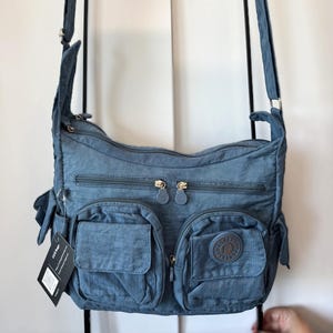 May include: A blue crossbody bag with multiple zippered compartments and pockets. The bag has a textured, crinkled appearance and features two front pockets with a circular logo. The bag is suspended from a black metal frame.