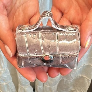 Op de afbeelding: Miniatuur zilverkleurige handtas met een getextureerd, krokodilachtig patroon. De kleine portemonnee heeft een handvat aan de bovenkant, een zilveren sluiting en decoratieve zilveren accenten. De tas wordt in open handen gehouden, wat de compacte afmetingen en het ontwerp benadrukt.