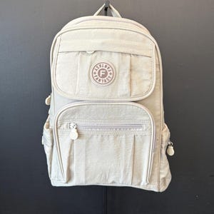May include: A light beige backpack with multiple zippered compartments and a top handle. The backpack features a round patch with the word "FANTASY" and the letter "F" in the center. The bag is hanging against a dark background.
