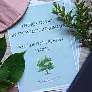 May include: A stack of printed guides titled "Things to Do in the Woods in Summer: A Guide for Creative People." The guides are light blue with green tree illustrations and text. Green leaves and a pink circular object are arranged around the guides.