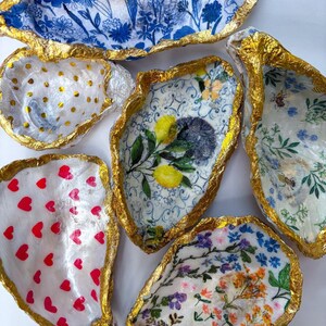May include: A collection of decorative oyster shell dishes with gold leaf edges. The shells feature various designs, including floral patterns, polka dots, and red hearts. These unique dishes are perfect for holding jewelry or small items.
