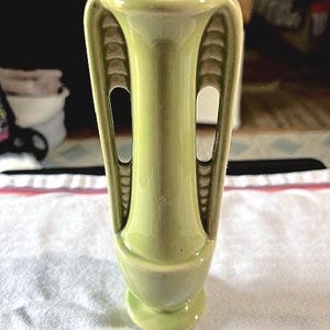 May include: A light green ceramic vase with a rounded base and a flared top. The vase has two vertical handles with cut-out sections. The vase is approximately 8 inches tall.