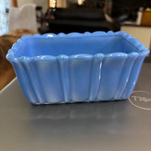 May include: A light blue, rectangular glass dish with a scalloped rim. The dish has vertical, fluted sides and a slightly rounded base. The color is a soft, opaque blue, and the dish appears to be vintage.