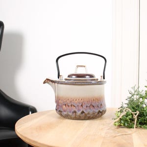 May include: A ceramic teapot with a black handle and lid. The teapot has a cream-colored top and a bottom with a textured, multi-colored design in shades of brown, purple, and beige. It sits on a round wooden table.