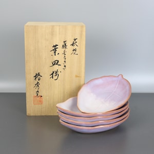 Puede incluir: Un conjunto de seis platos de cerámica con forma de hoja, en tonos lavanda y blanco. Los platos están apilados, mostrando su delicada forma y diseño de hoja veteada. Una caja de madera con caligrafía japonesa se encuentra en el fondo.