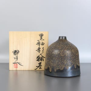 Könnte beinhalten: Eine Keramikvase mit einer strukturierten braunen und schwarzen Glasur und einem dunklen Sockel. Die Vase hat eine abgerundete Form mit schmalem Hals. Links von der Vase befindet sich eine Holzkiste mit japanischer Kalligraphie.