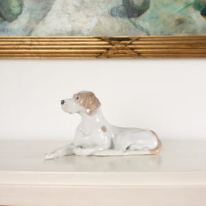 Puede incluir: Figura de porcelana de un perro en posición de descanso. El perro es principalmente blanco con marcas marrones en la cabeza y el cuerpo. La figura se exhibe sobre una superficie blanca, con una pintura en un marco dorado al fondo.