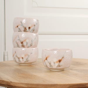 Puede incluir: Cuatro cuencos de cerámica rosa con una superficie texturizada y un diseño moteado en marrón y blanco. Un cuenco está separado, mientras que los otros tres están apilados. Los cuencos están sobre una mesa de madera clara.