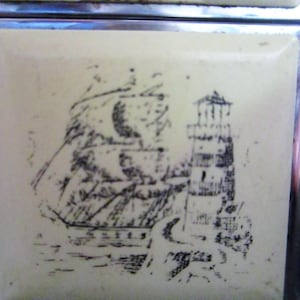 May include: A vintage lighter with a white background and a black and white image of a ship and a lighthouse.