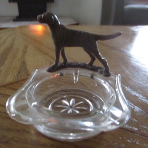 Peut inclure: Un cendrier en verre transparent avec une figurine de chien brune sur le dessus. Le cendrier a un motif décoratif sur le fond.