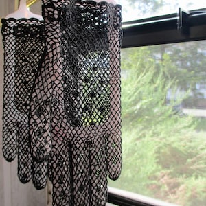 May include: A pair of black lace gloves hanging on a hanger. The gloves are made of a delicate, intricate lace pattern. The gloves are hanging in front of a window with a screen.